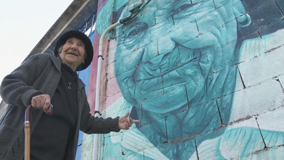 Remedios Cabanelas posa diante do mural que protagoniza no centro da súa aldea de Puxedo